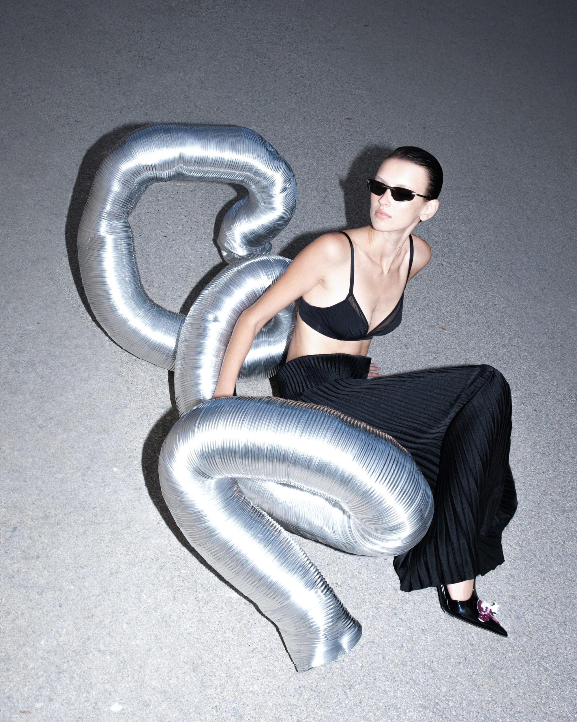 Model in black bralette and pleated trousers leaning against a large metallic inflatable tube on a concrete background.