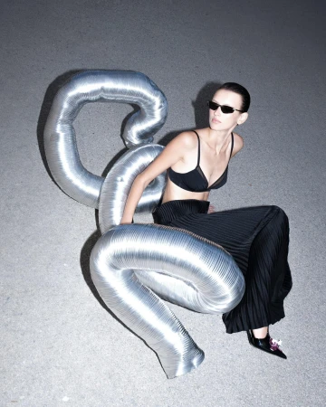 Model in black bralette and pleated trousers leaning against a large metallic inflatable tube on a concrete background.