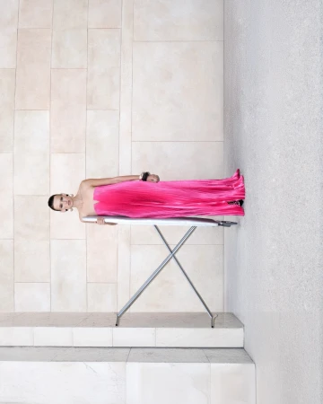 Model wearing a vibrant pink pleated strapless dress leaning against an ironing board outdoors