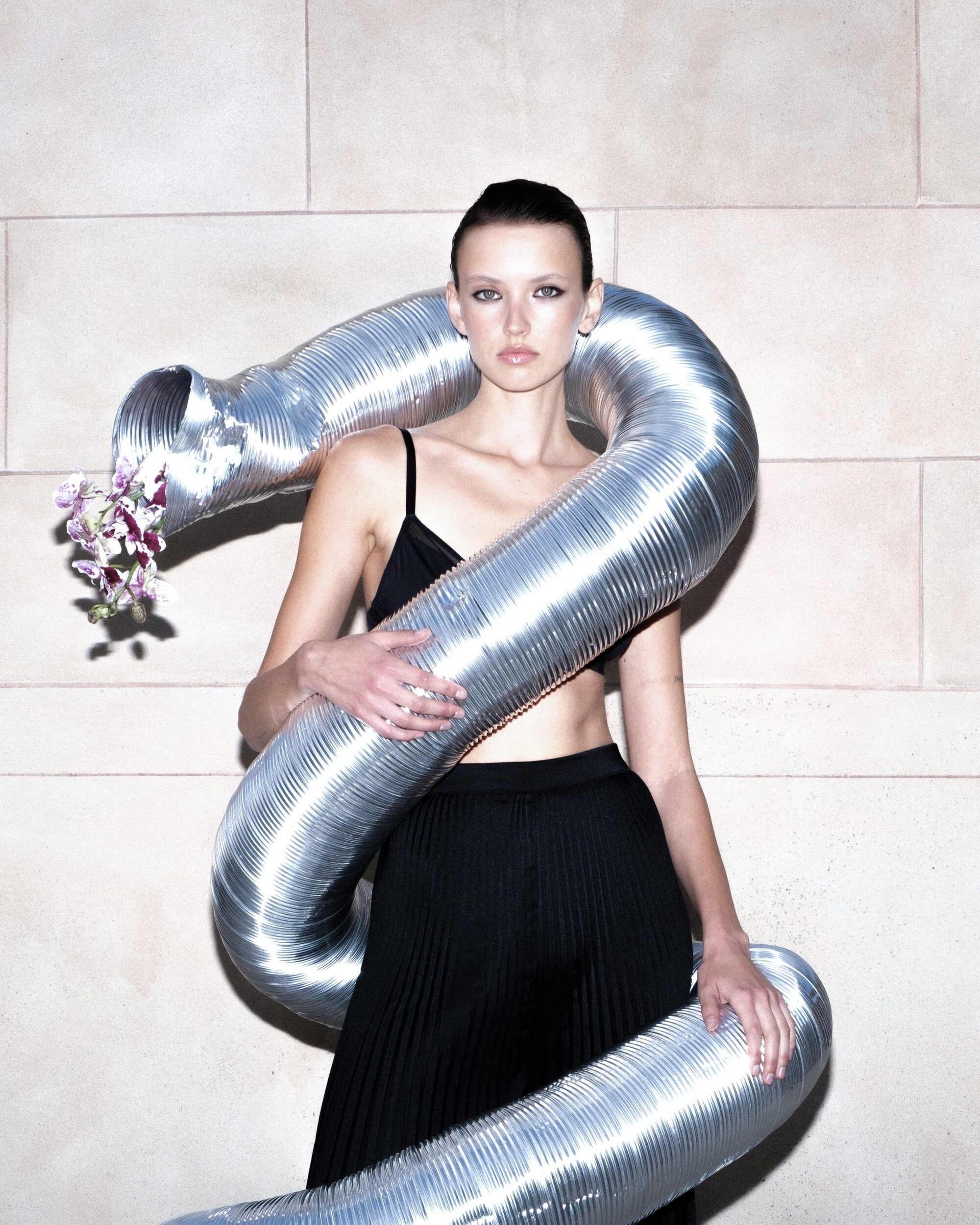 Model in black wearing a winding metallic duct tube with orchids against a stone block wall