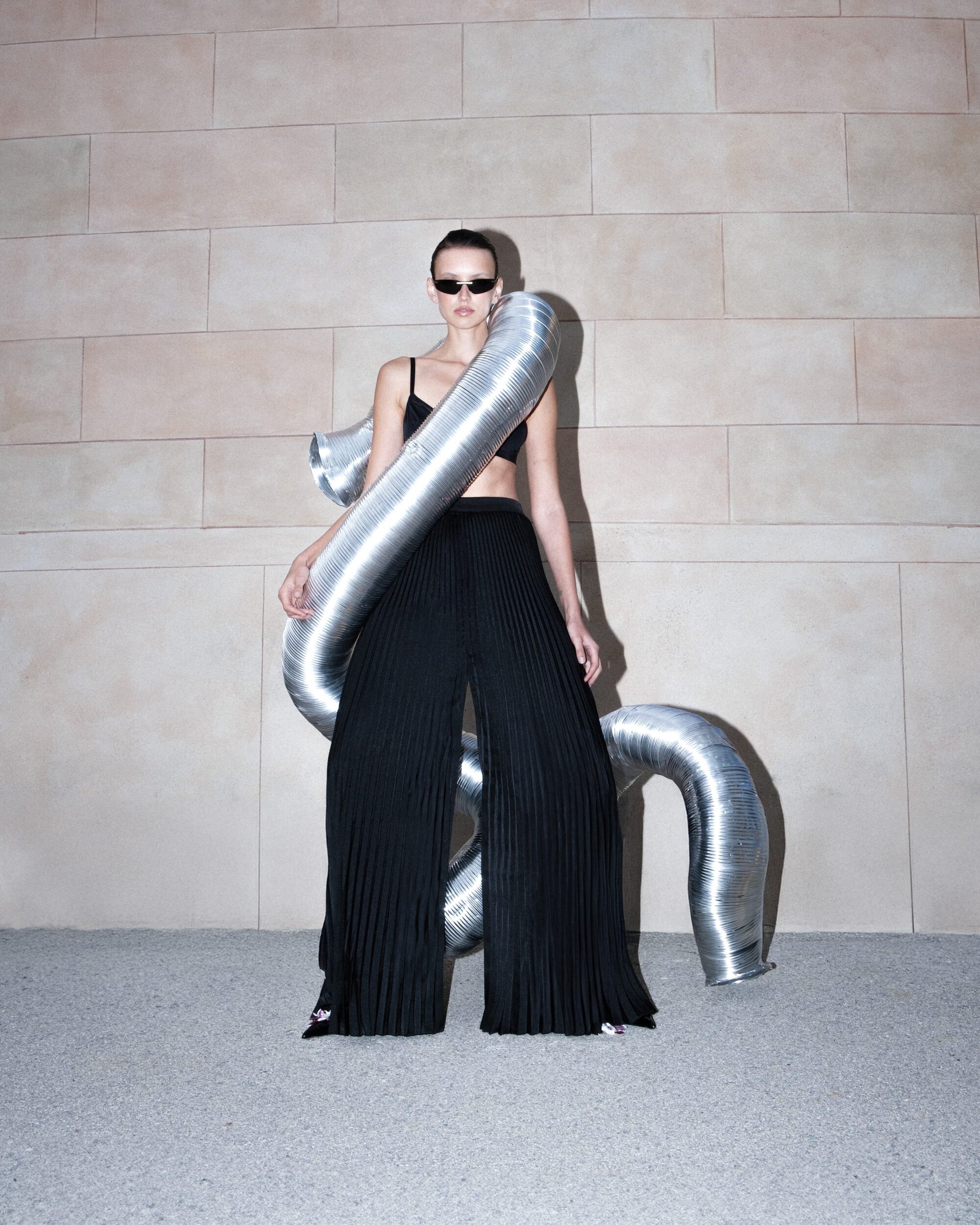 Model in black top and pleated trousers posed with a metallic flexible duct against a stone block wall