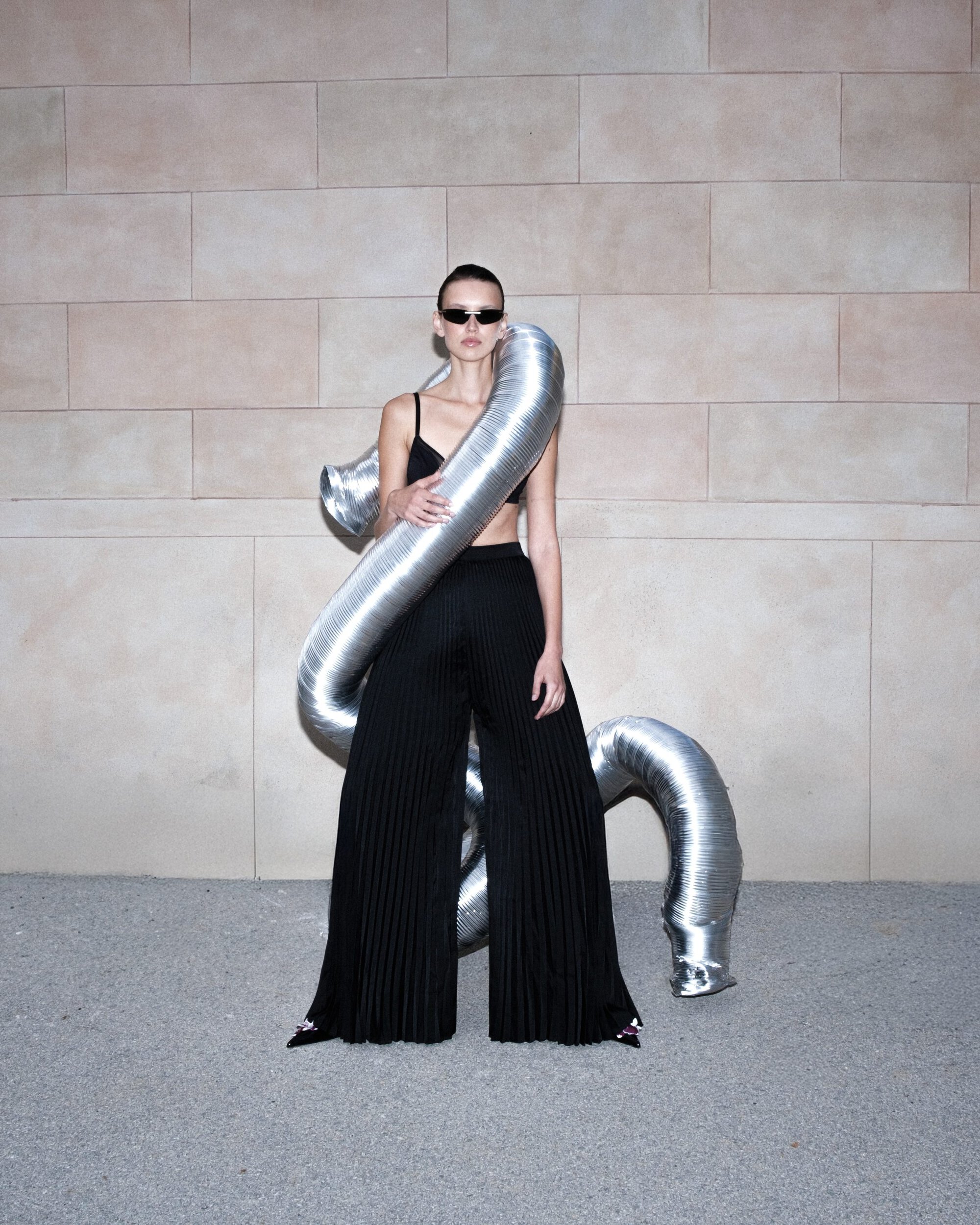 Model wearing dark sunglasses and black pleated trousers with a metallic silver flexible duct wrapped around her torso.