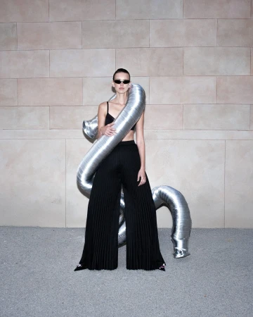 Model wearing dark sunglasses and black pleated trousers with a metallic silver flexible duct wrapped around her torso.