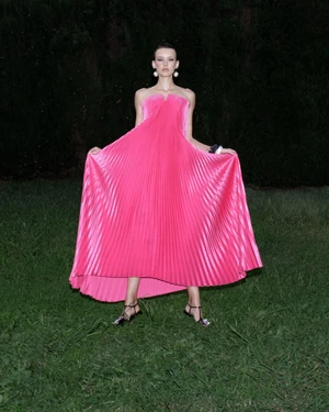 Model wearing a vibrant pink pleated strapless dress standing outdoors on a lawn at night for L'idee Woman Fall 2024 ad campaign.