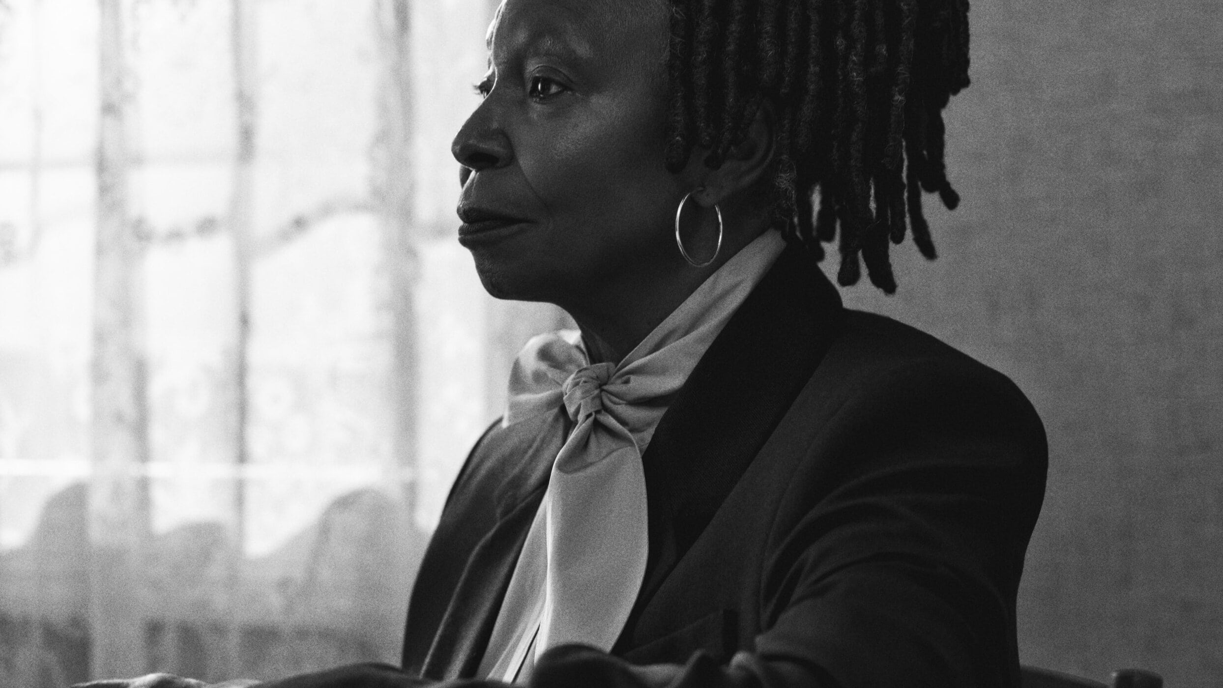 Monochrome portrait of Whoopi Goldberg wearing a dark blazer and large pussy-bow blouse for Ami Fall 2024 ad campaign.