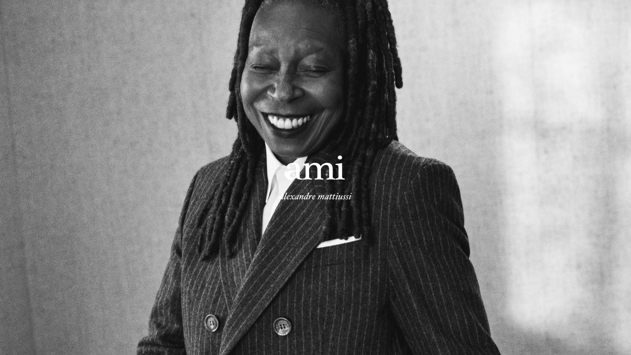 Whoopi Goldberg wearing a dark pinstripe suit jacket smiling in a black and white portrait for Ami Fall 2024 campaign