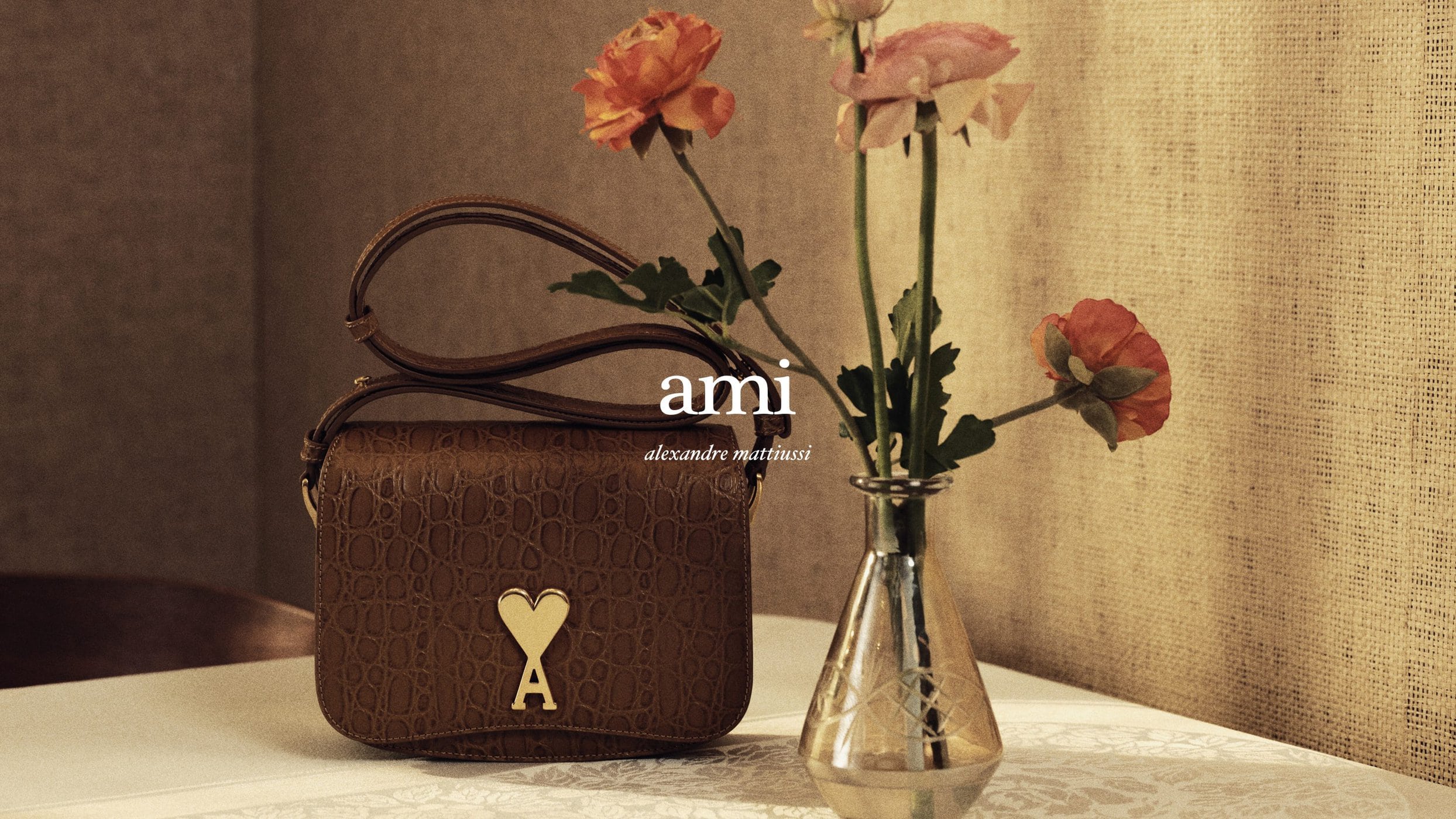 Brown leather handbag with gold heart emblem resting on a white table next to a glass vase with orange flowers