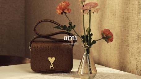 Brown leather handbag with gold heart emblem resting on a white table next to a glass vase with orange flowers