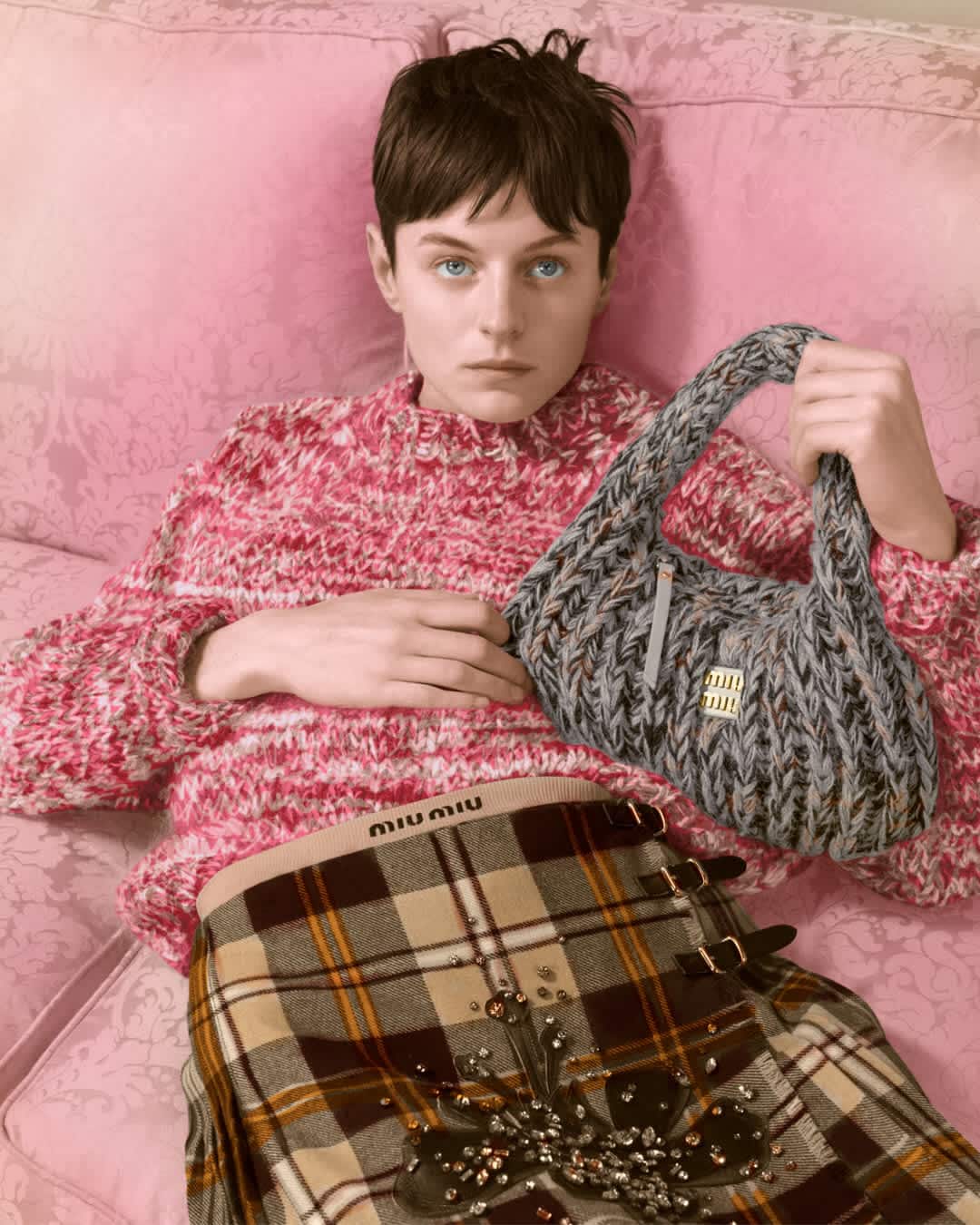 Model Emma Corrin holding a grey knit handbag while seated on a pink couch for Miu Miu Upcycled Holiday 2024 campaign