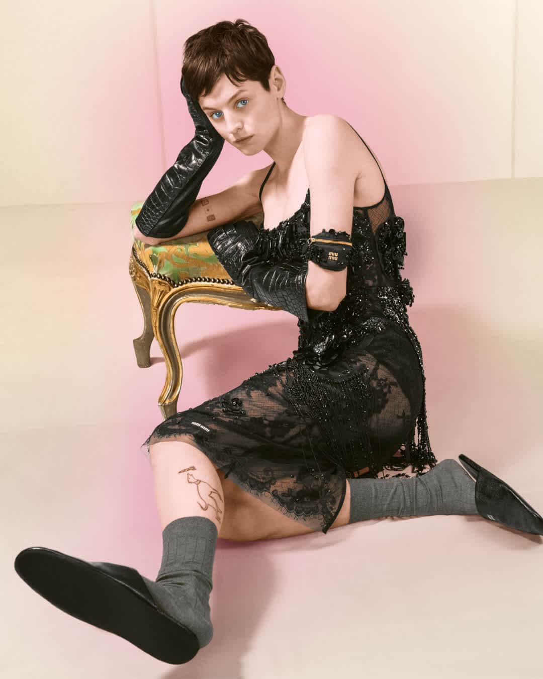 Model Emma Corrin seated on an ornate stool wearing a black lace slip dress and long leather gloves for Miu Miu