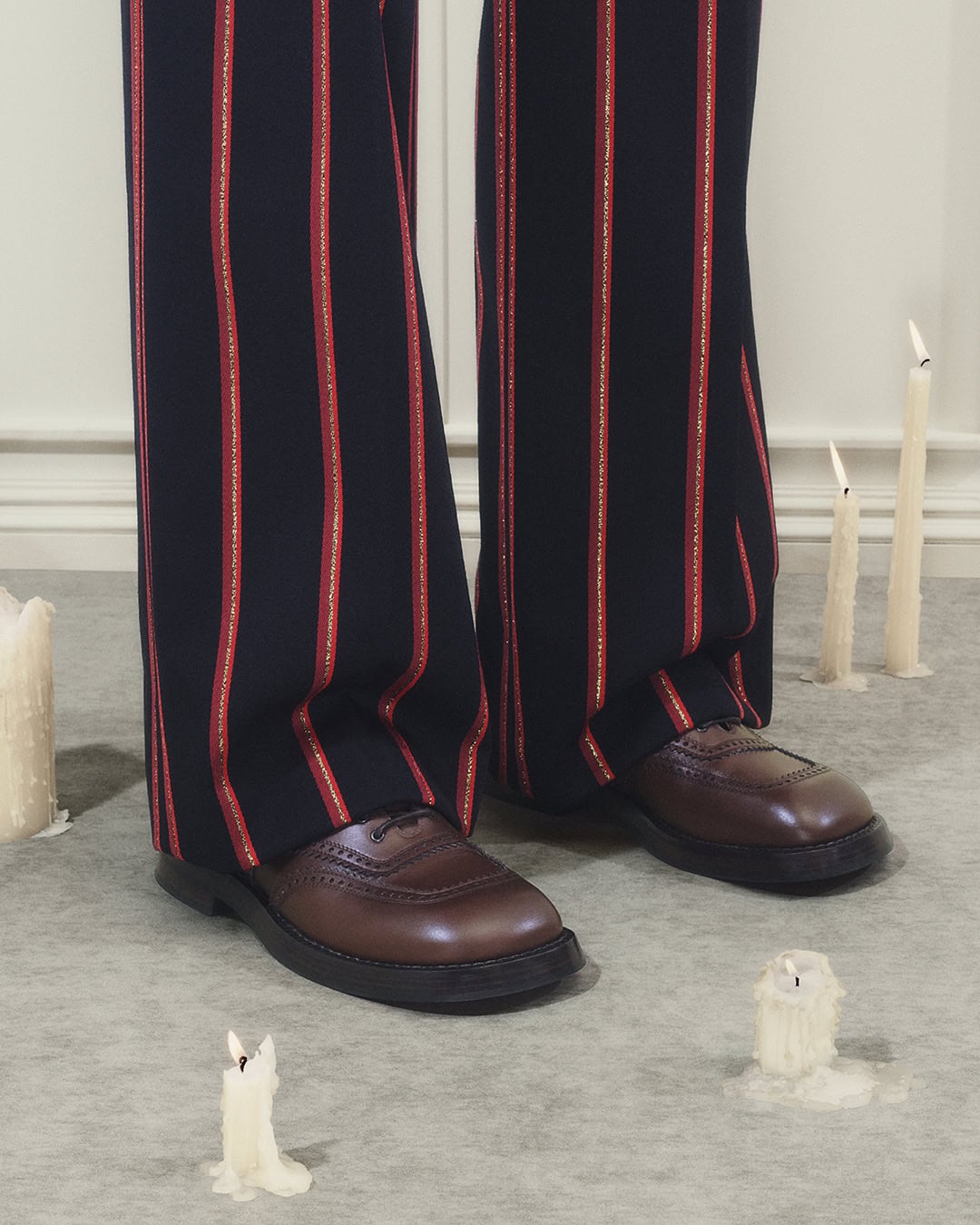 Close-up of trousers with red stripes and brown leather shoes surrounded by lit candles on grey floor