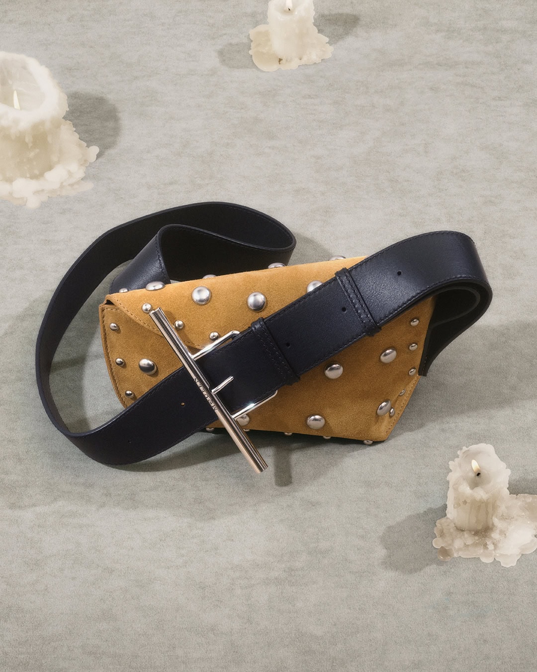 Tan suede studded handbag with a thick black leather strap resting on a textured surface surrounded by melting candles.