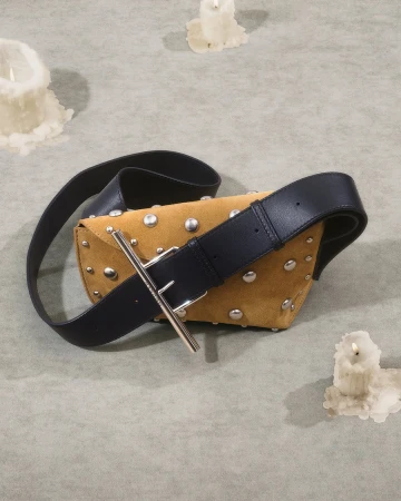 Tan suede studded handbag with a thick black leather strap resting on a textured surface surrounded by melting candles.