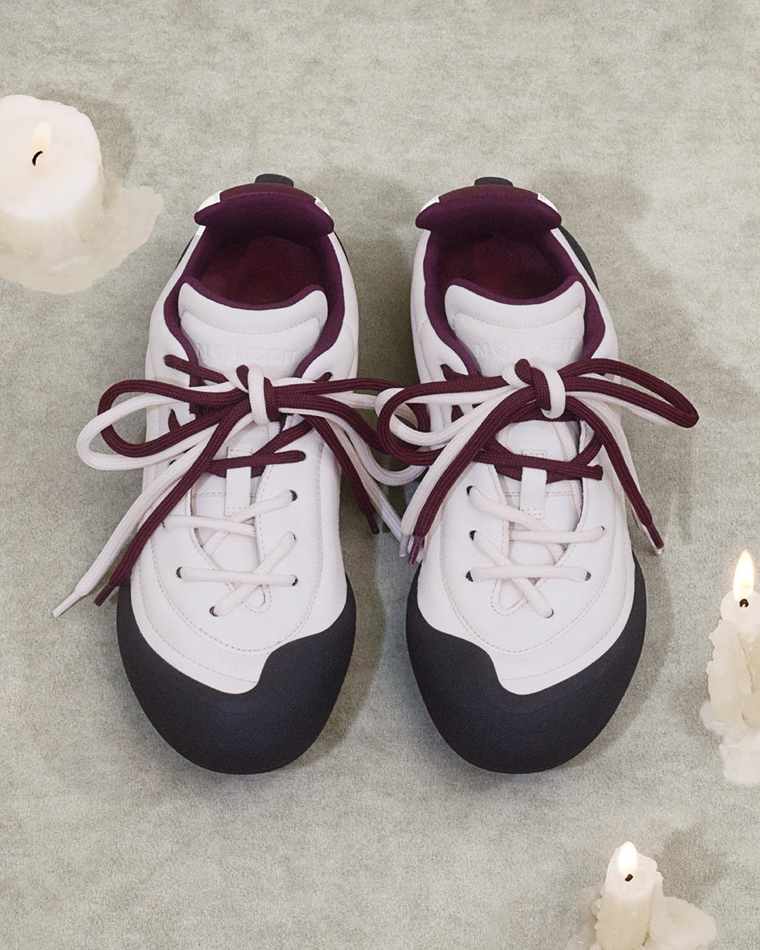 Top-down view of white sneakers with dark toes and burgundy laces placed on a textured grey background with burning candles.