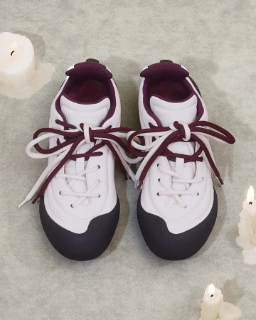 Top-down view of white sneakers with dark toes and burgundy laces placed on a textured grey background with burning candles.