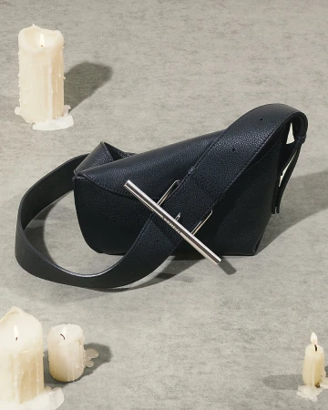 Black leather shoulder bag with a silver metal bar closure resting on a textured grey surface surrounded by burning lit white candles.