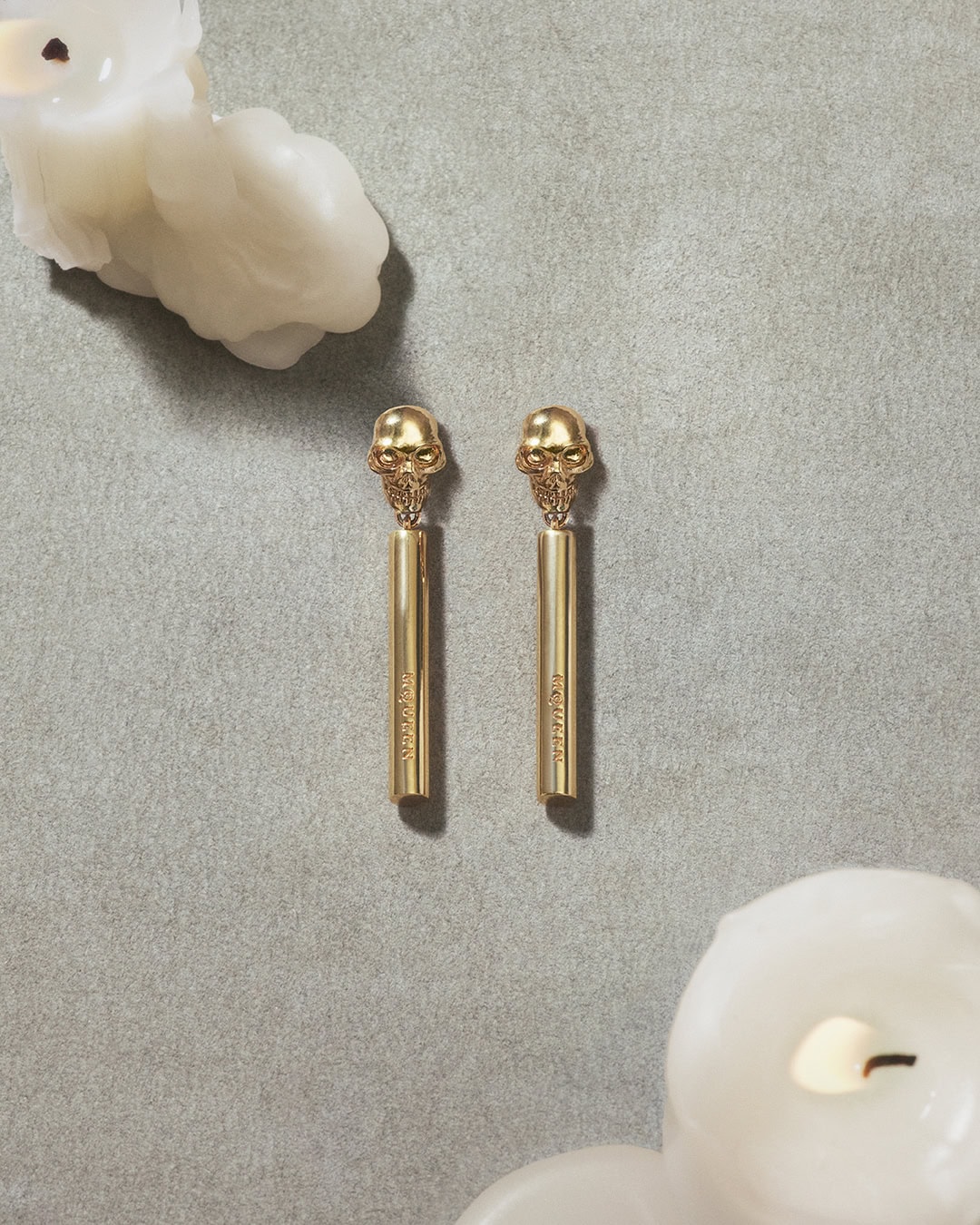 Pair of gold skull earrings displayed on a grey textured surface with melting white candles in opposite corners.