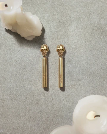 Pair of gold skull earrings displayed on a grey textured surface with melting white candles in opposite corners.