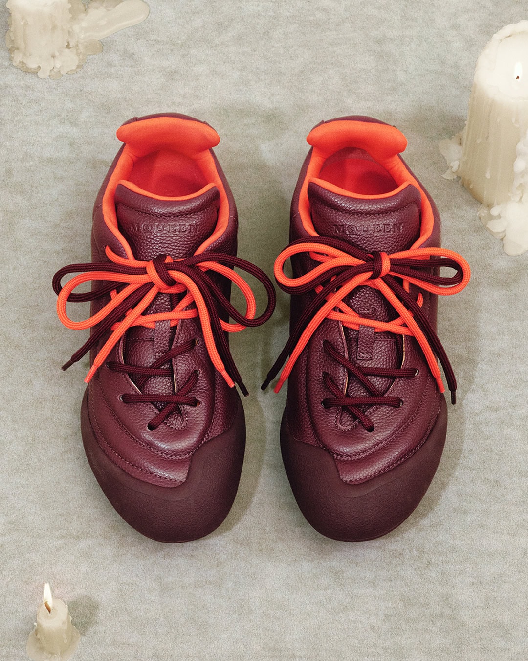 Top down view of a pair of burgundy and orange leather sneakers resting on a textured grey surface with lit candles