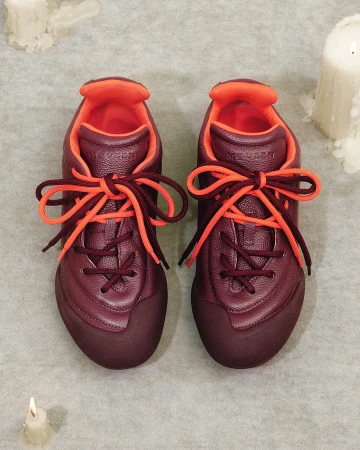Top down view of a pair of burgundy and orange leather sneakers resting on a textured grey surface with lit candles