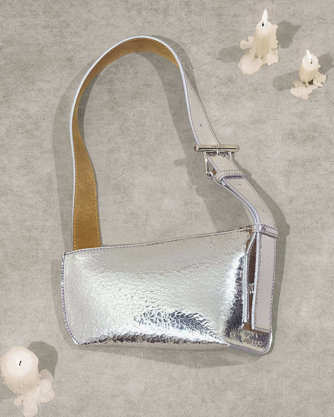 Silver metallic leather shoulder bag with a buckle strap on a grey textured background with three white candles.
