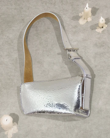Silver metallic leather shoulder bag with a buckle strap on a grey textured background with three white candles.
