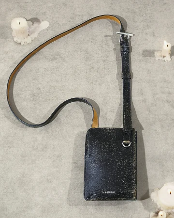Black leather crossbody bag with a thin strap resting on a textured grey surface next to melting white candles