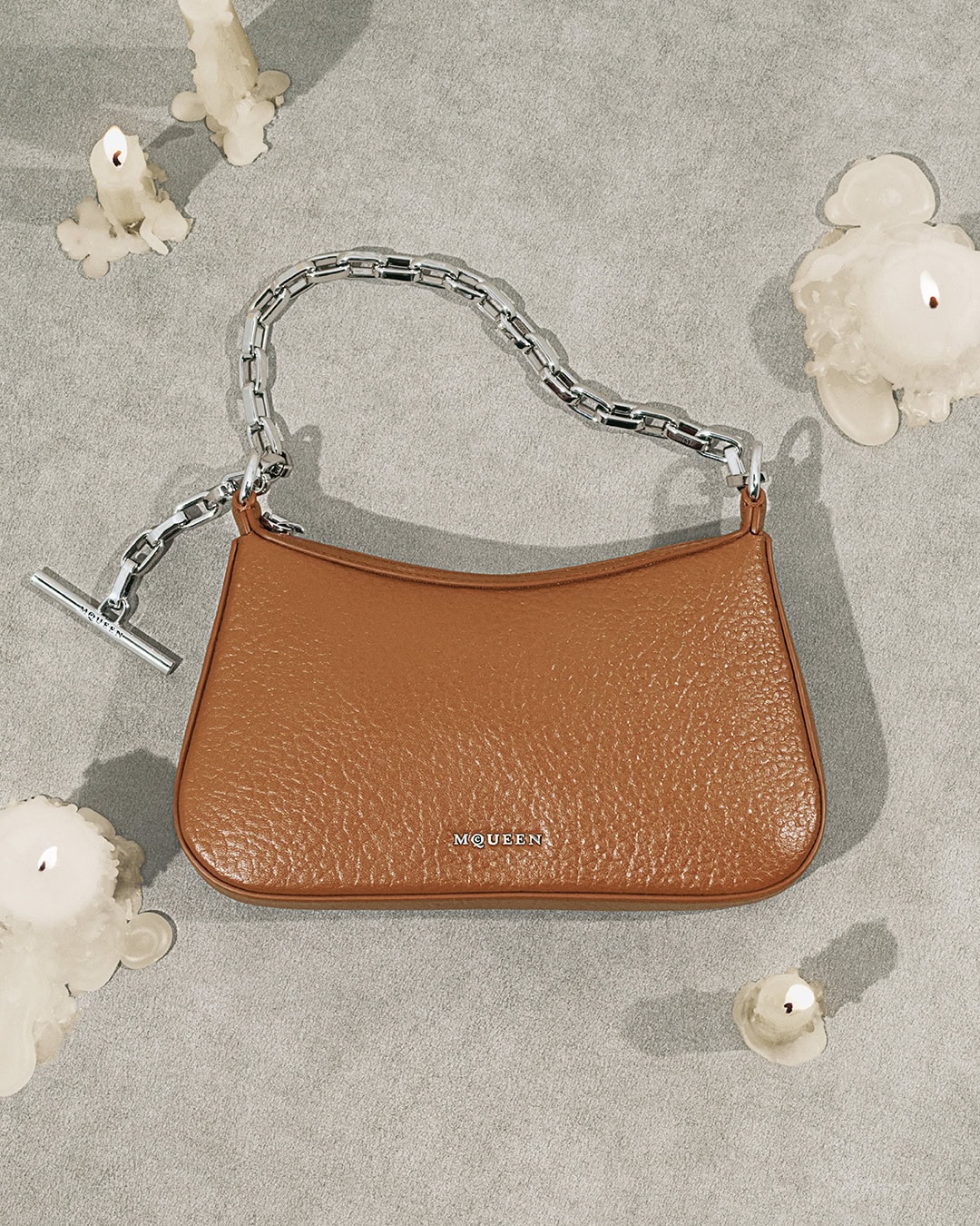 Brown leather handbag with a thick silver chain strap resting on a textured gray background surrounded by melting candles