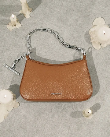 Brown leather handbag with a thick silver chain strap resting on a textured gray background surrounded by melting candles