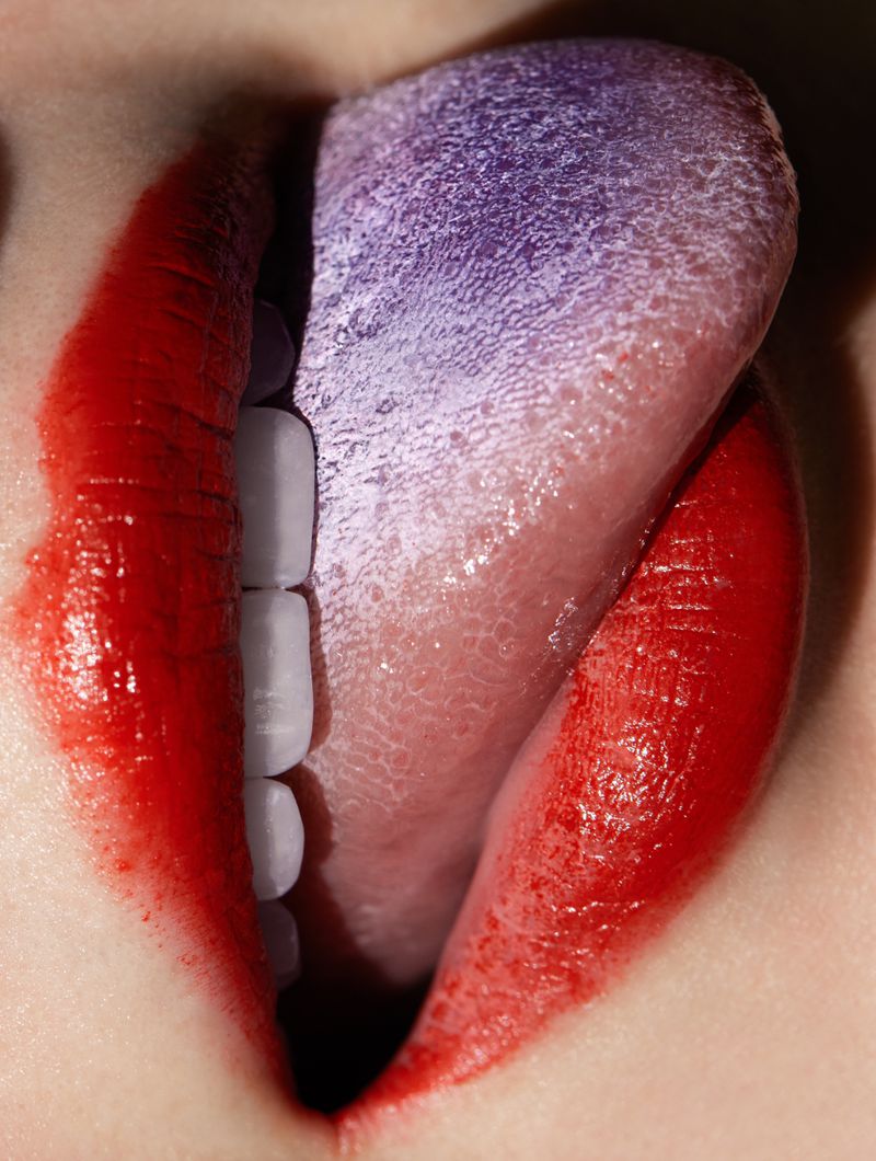Close-up of a person's mouth with red lipstick and visible teeth, tongue with purple tint, creating a bold and striking visual.