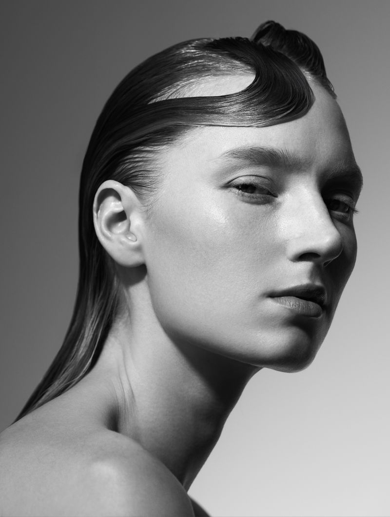 Monochrome portrait of a young woman with stylized slicked-back hair, minimal makeup, and neutral expression in a studio setting.