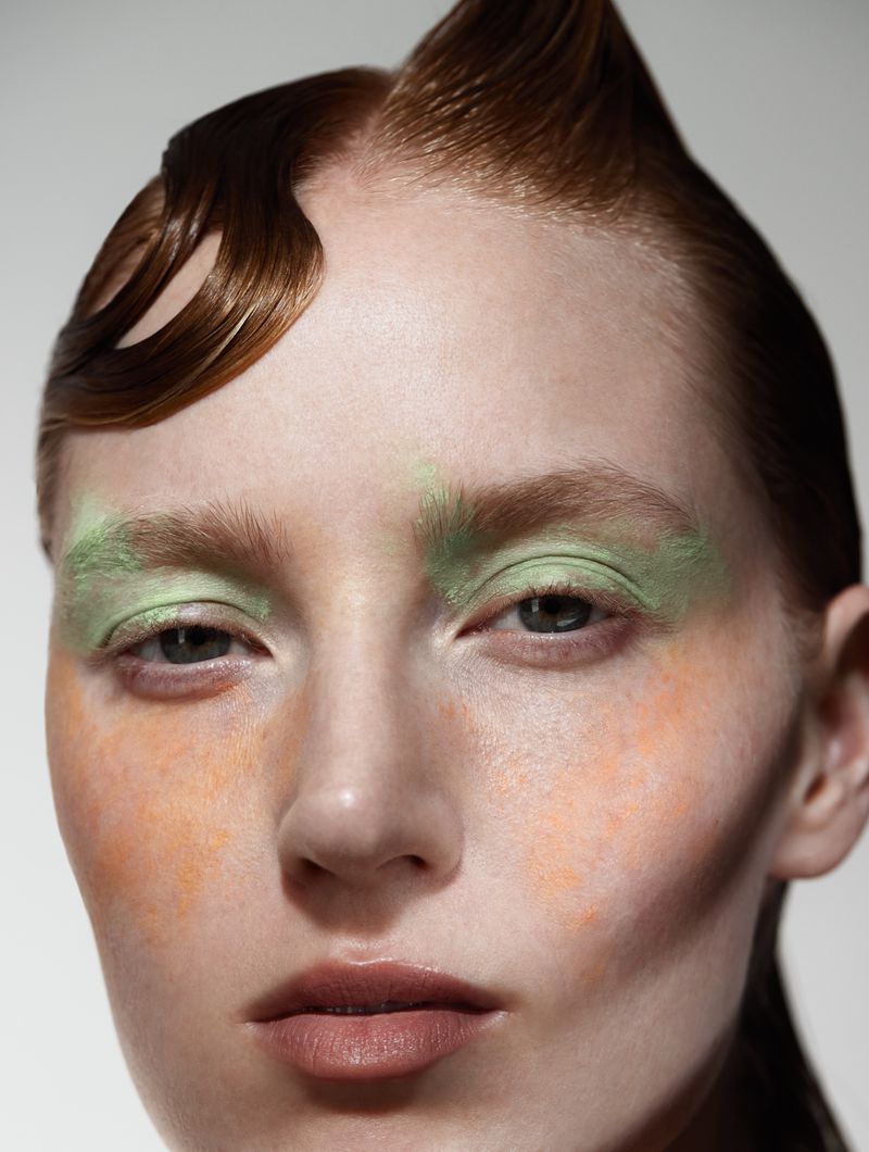Close-up portrait of a fair-skinned woman with elaborate red hair, wearing mint green eyeshadow and orange blush on a grey background.