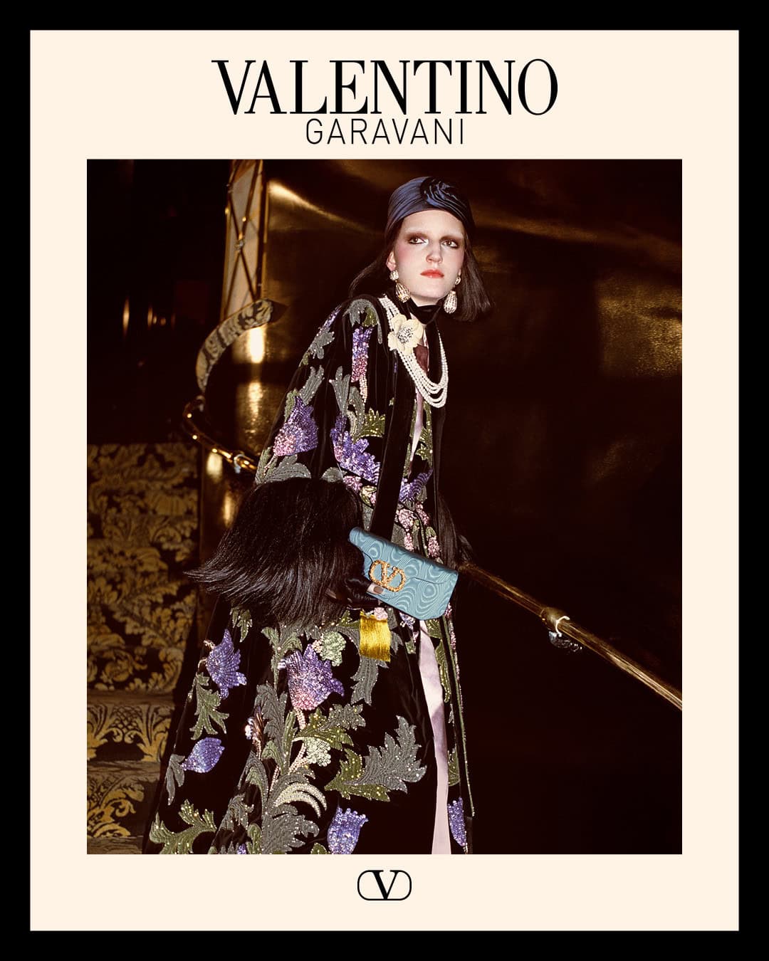 Model in embellished dark coat and headpiece holding a blue handbag on a grand staircase for Valentino