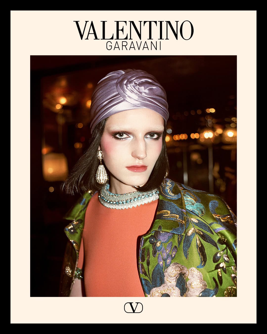 Model wearing a purple satin turban and green floral brocade jacket with ornate drop earrings for Valentino Holiday 2024 campaign