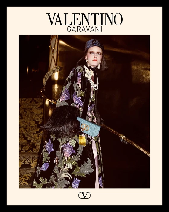 Model in embellished dark coat and headpiece holding a blue handbag on a grand staircase for Valentino