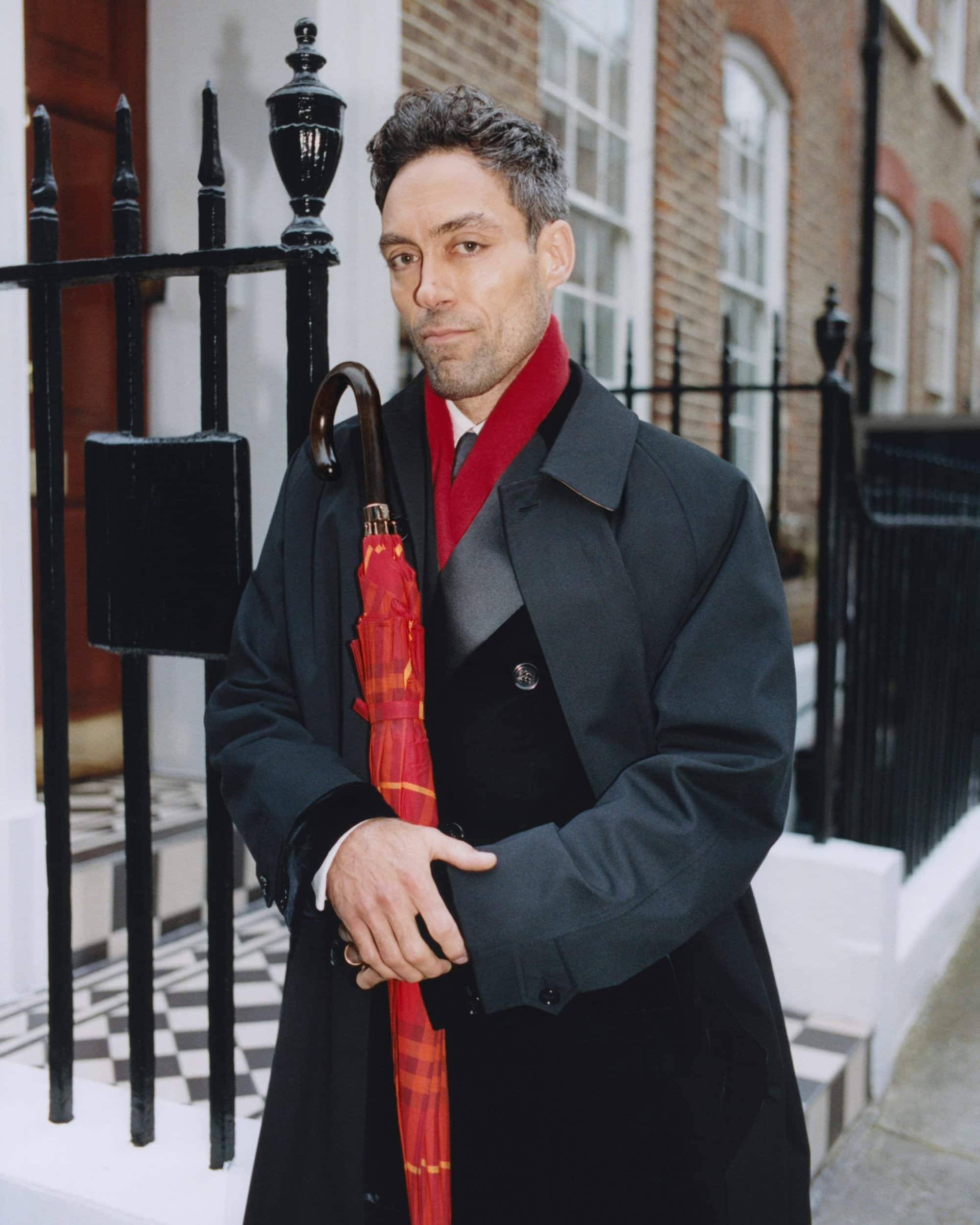 Man wearing a black coat and red scarf holding a red umbrella standing outside a London building with black iron railings