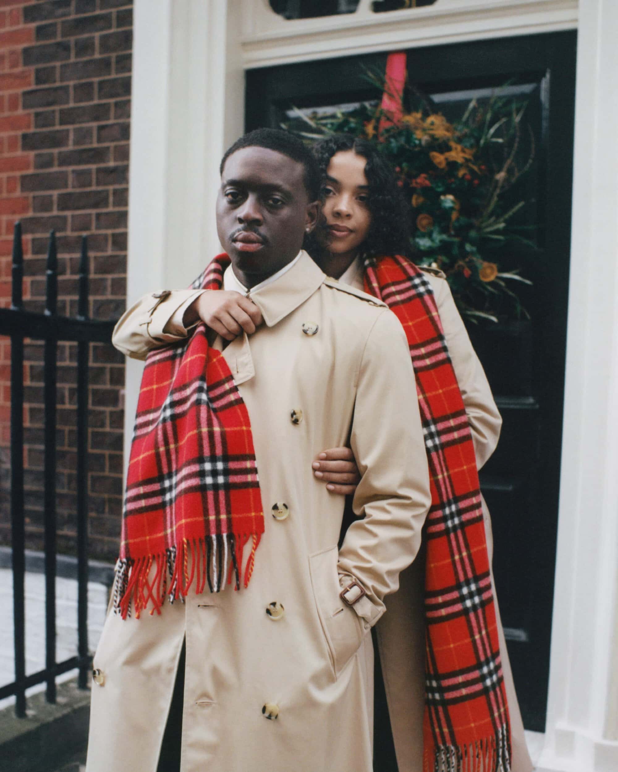 Two models wearing beige trench coats and red plaid scarves standing outside a brick building in London.