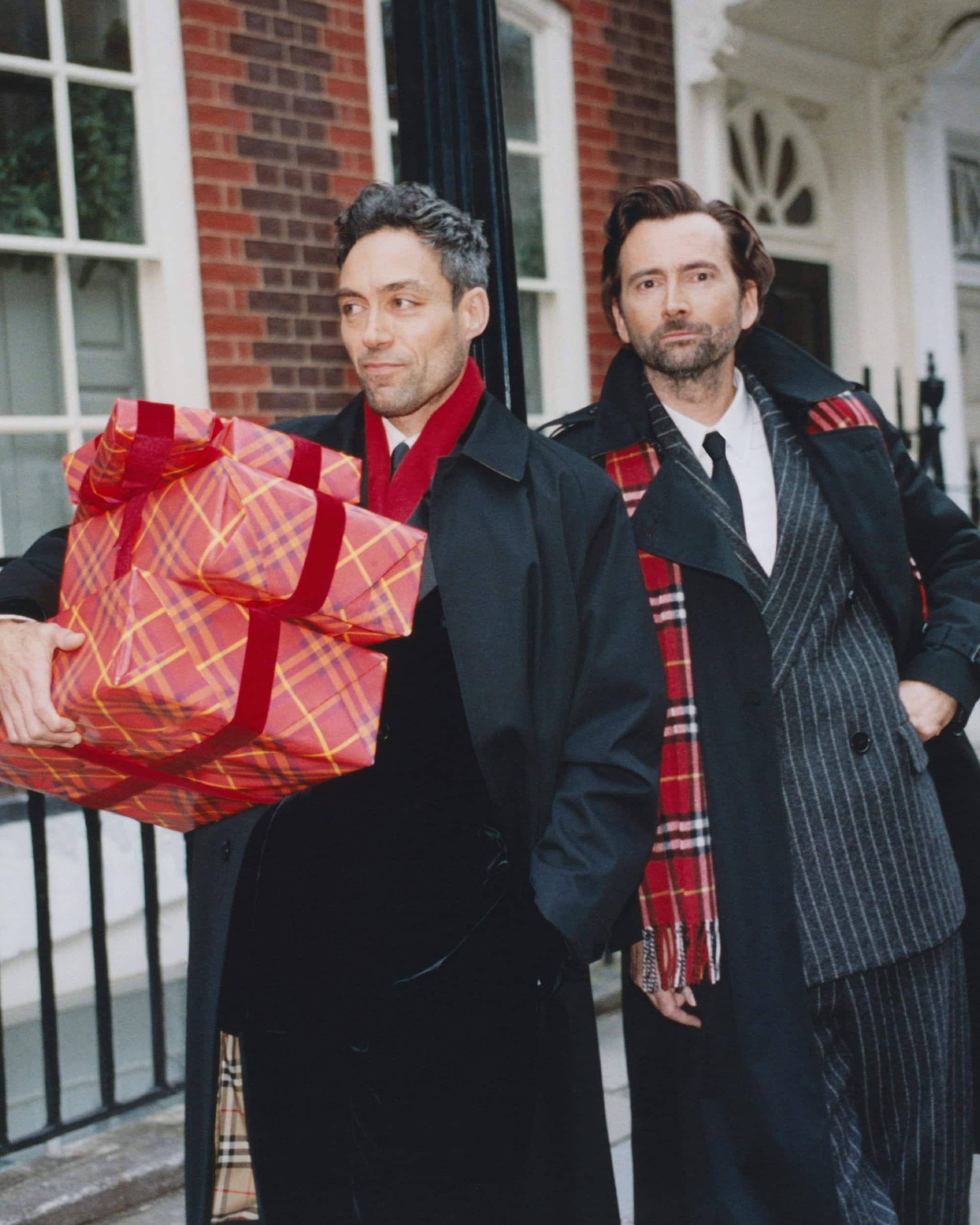 Two men wearing dark coats and plaid scarves carrying wrapped holiday gifts outside a brick building