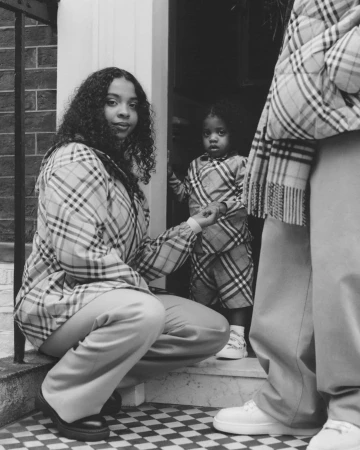 Black and white portrait of a woman crouching next to a child in a doorway wearing plaid outerwear for Burberry Holiday 2024
