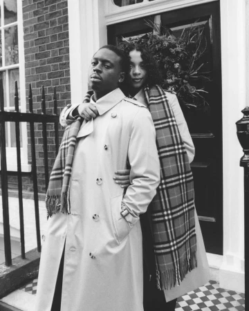 Black and white portrait of two models wearing trench coats and plaid scarves in front of a brick exterior.