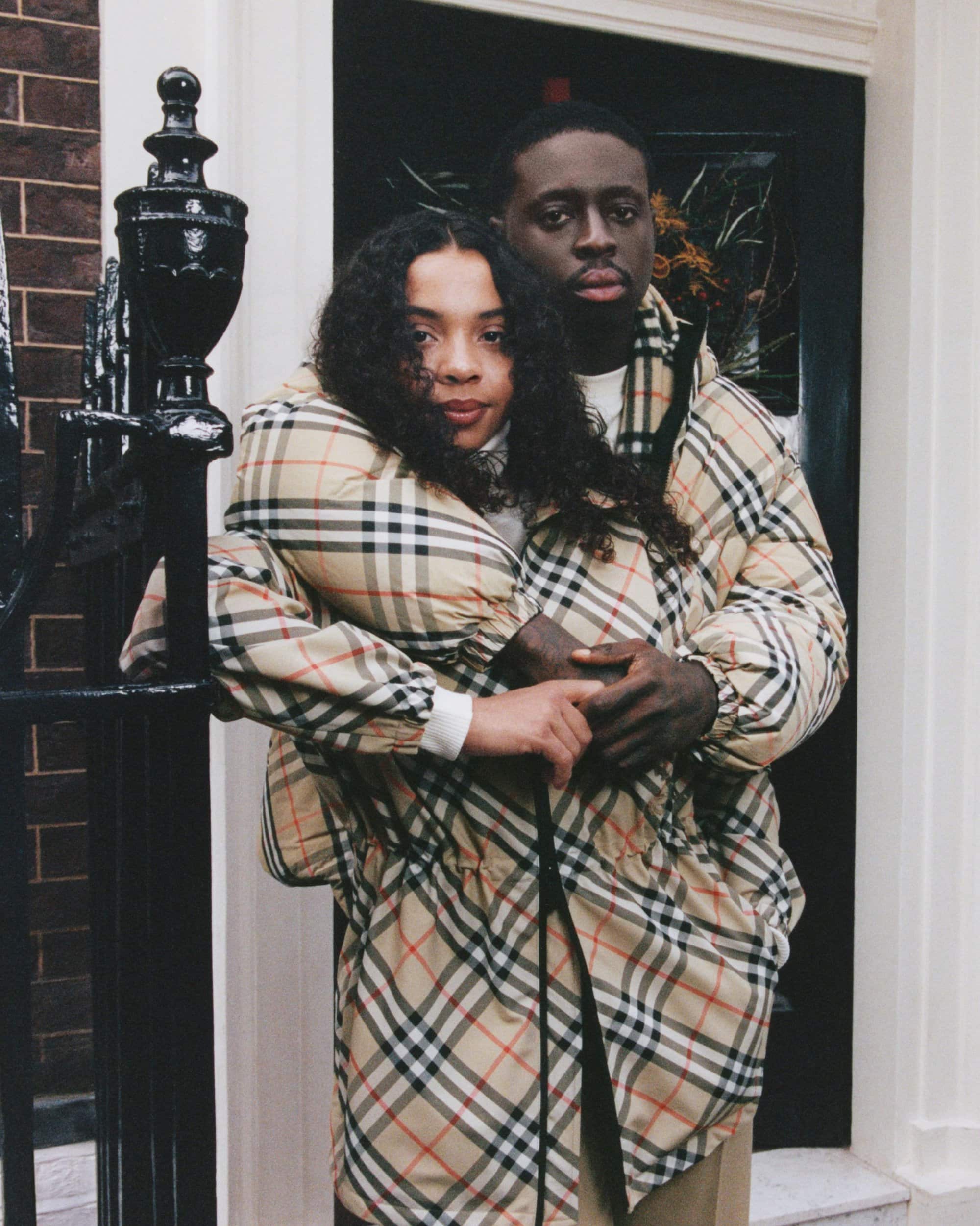 A man and woman wearing matching plaid jackets embracing outdoors near a dark doorway and brick wall