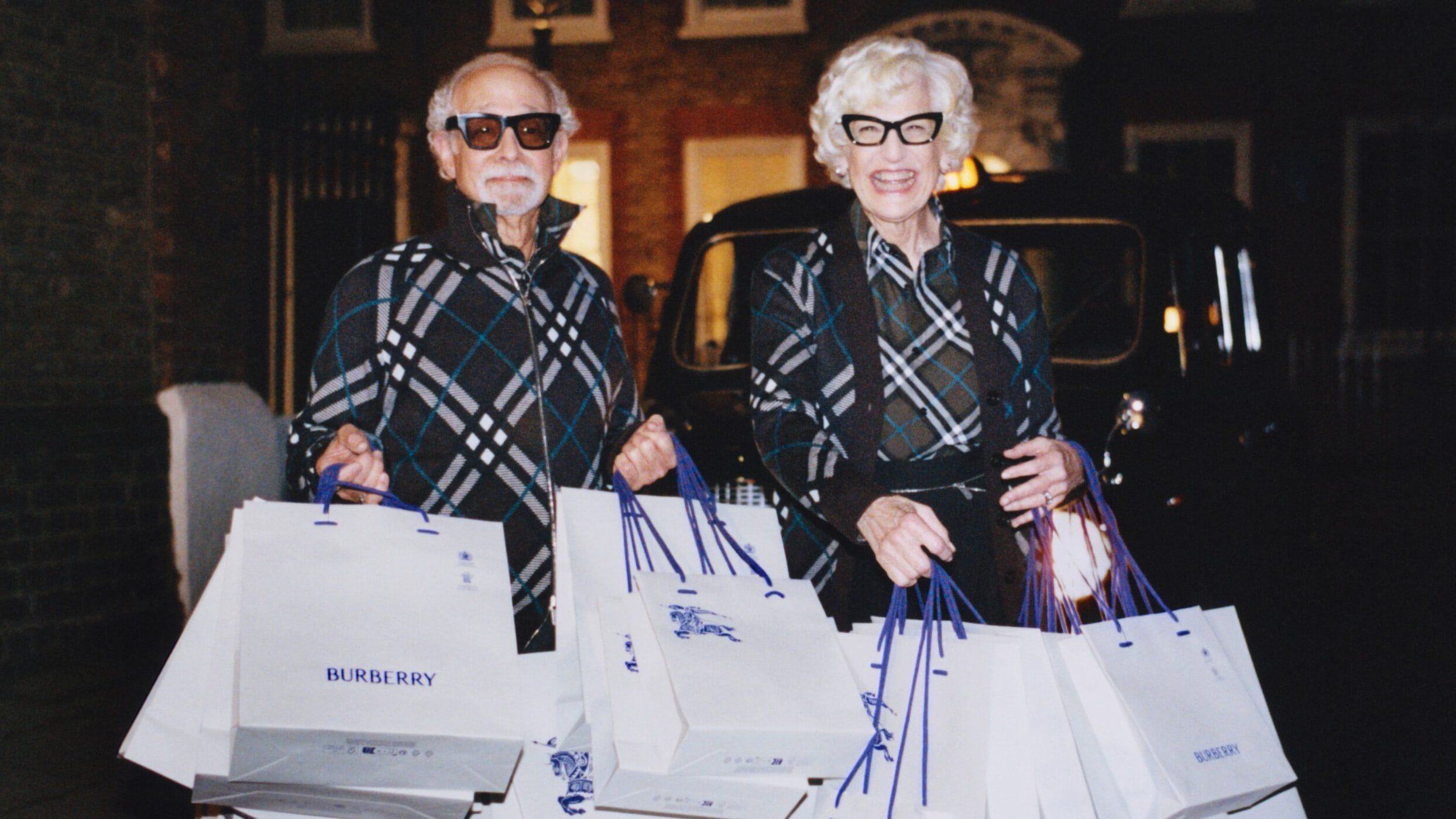 An elderly couple in patterned zip-up jackets holding multiple white shopping bags with blue handles outdoors at night.
