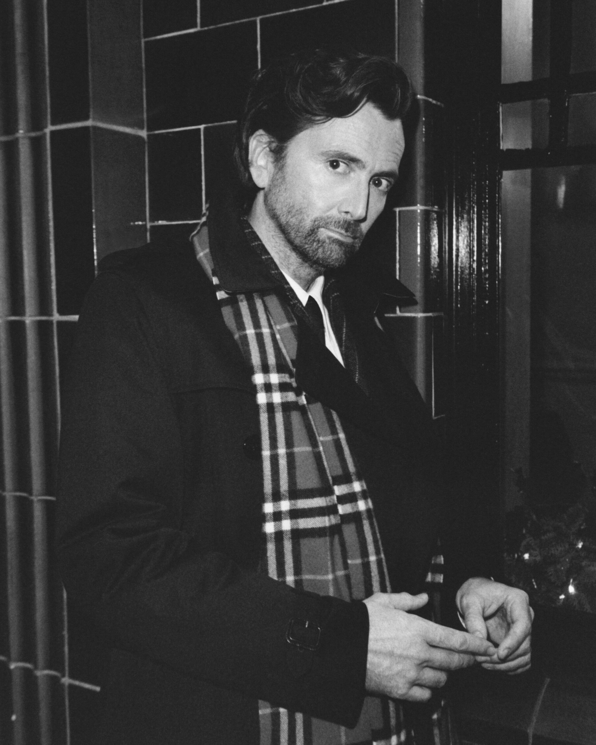David Tennant wearing a dark coat and plaid scarf standing indoors against a dark tiled wall in a black and white portrait.