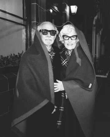 Older couple wrapped together in a heavy wool blanket wearing dark sunglasses and glasses outside at night