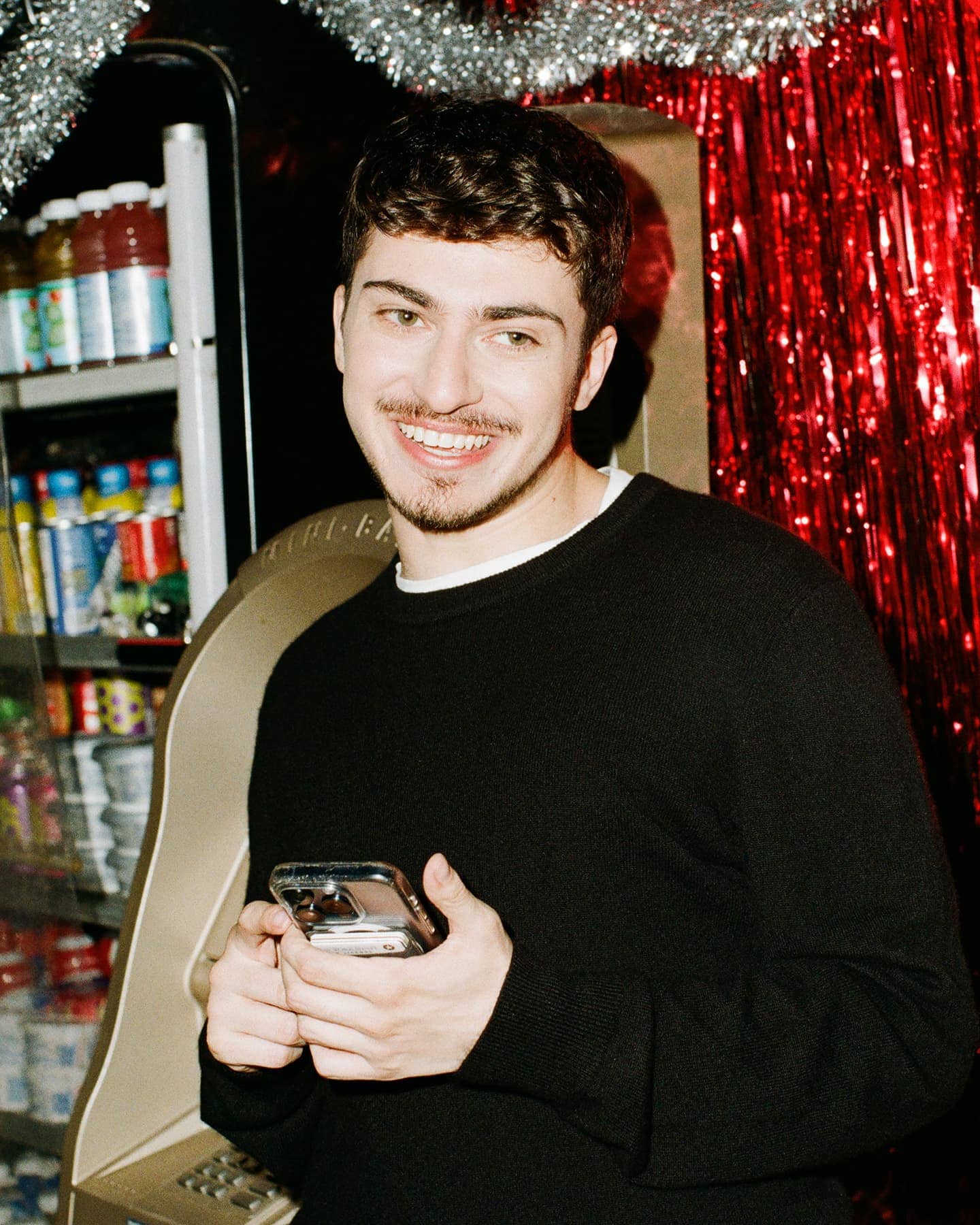 A smiling young man holding a smartphone in front of a red tinsel backdrop and vending machine for Michael Kors Holiday 2024 campaign.