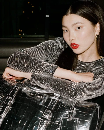 Model in sparkling silver top and red lipstick leaning over a metallic handbag in a car