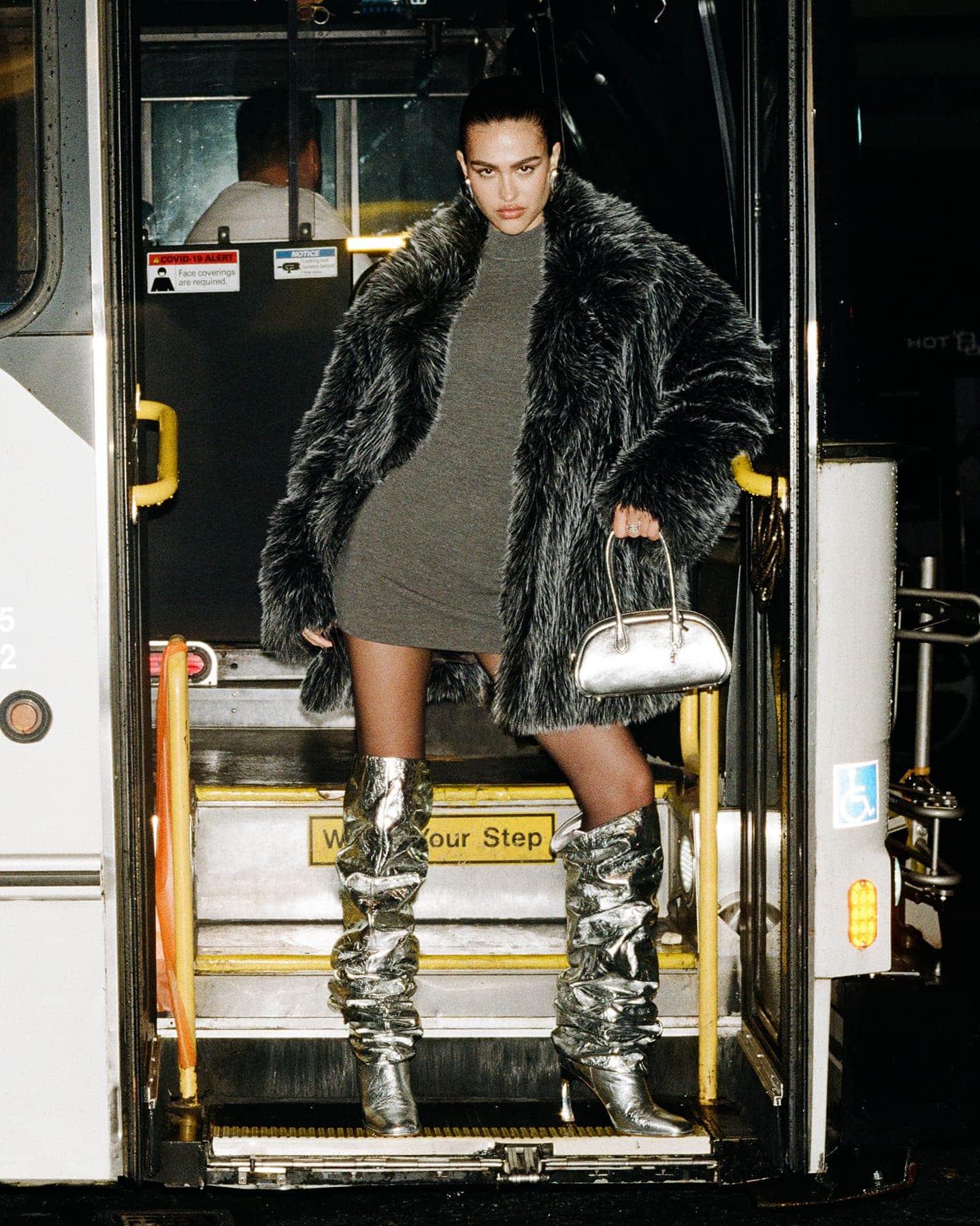 Model in a fuzzy dark coat and silver boots holding a silver handbag while stepping off a public transit bus.