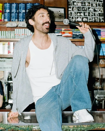 Man with curly dark hair and mustache holding a microphone while wearing a grey zip hoodie and blue jeans