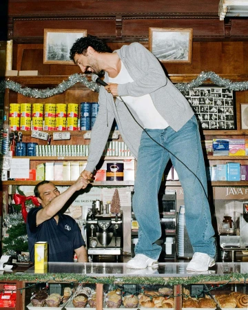 Man holding a microphone standing on a counter, shaking hands with a seated man in a shop