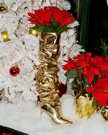 Metallic gold slouch boot filled with bright red poinsettia flowers standing in front of a white Christmas tree with gold ornaments.